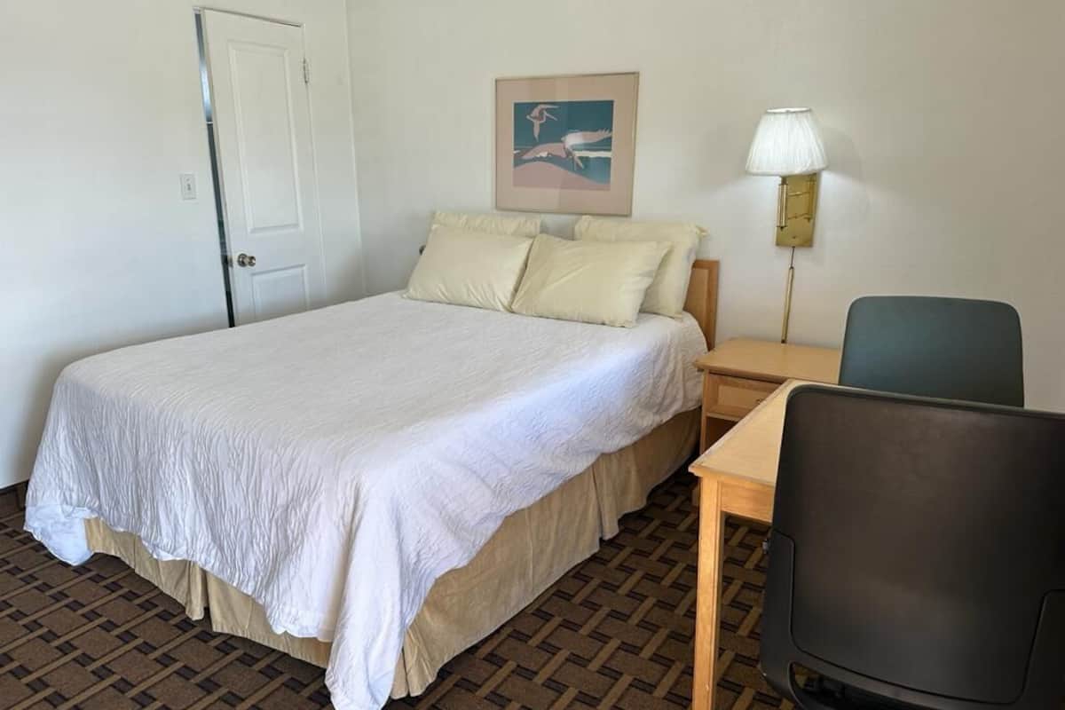 A hotel room with a bed, a desk, a chair, a lamp, and a door.