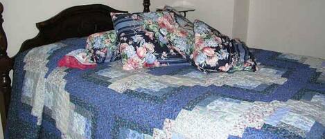 A bed with a floral quilt, a wooden headboard, and a white wall with a hanging lamp.