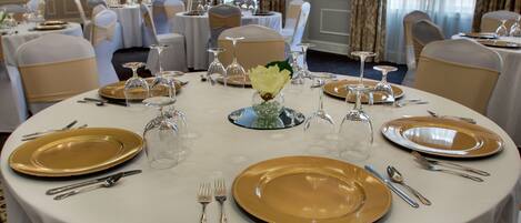 A banquet hall with round tables set for a formal event, complete with gold-rimmed plates, cutlery, and glassware.