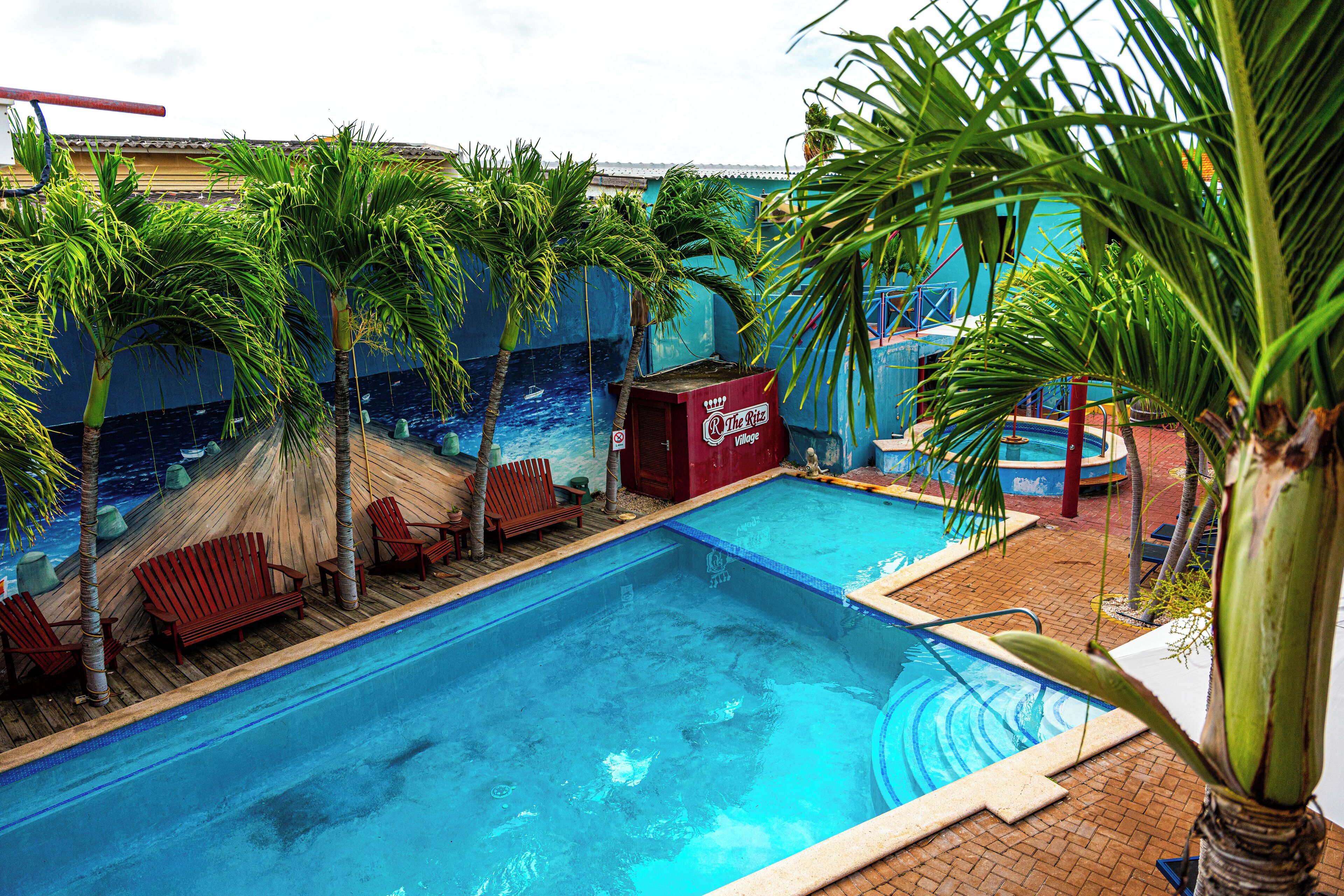 A swimming pool with palm trees, a red building labeled "The Ritz Village," and wooden deck chairs.