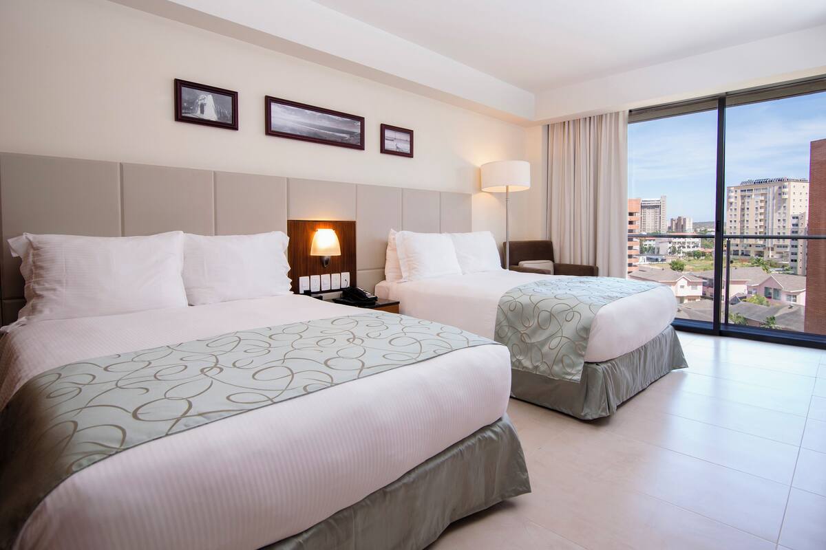 Une chambre d’hôtel avec deux lits, une grande fenêtre donnant sur la ville et une lampe fixée au mur.