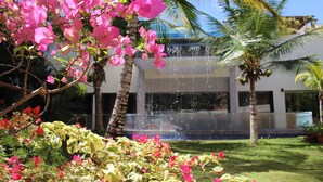 Un edificio con una fuente en primer plano, rodeado de palmeras y plantas con flores.