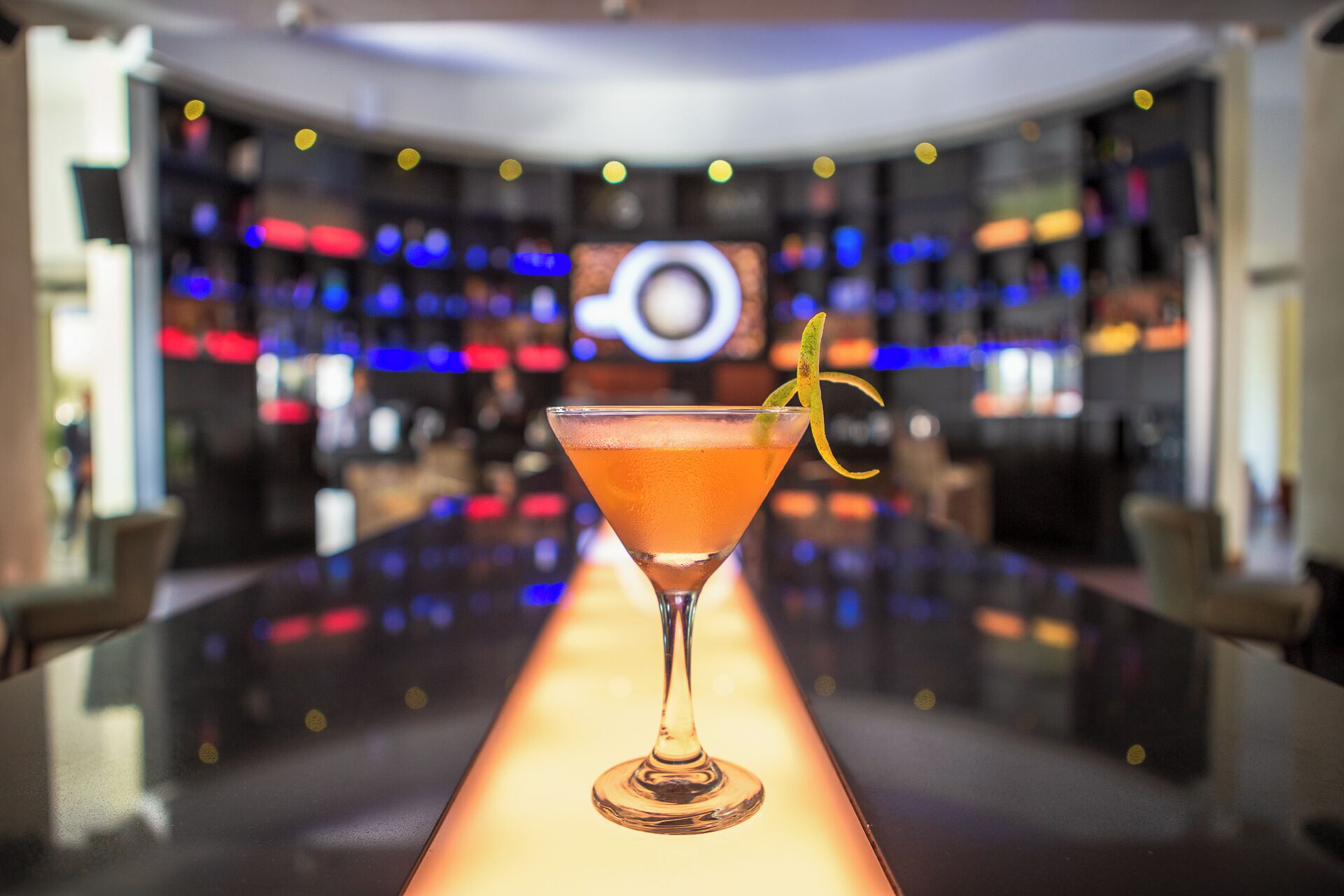 A bar with a cocktail in a martini glass, a yellow lemon peel garnish, and a blurred background with lights.