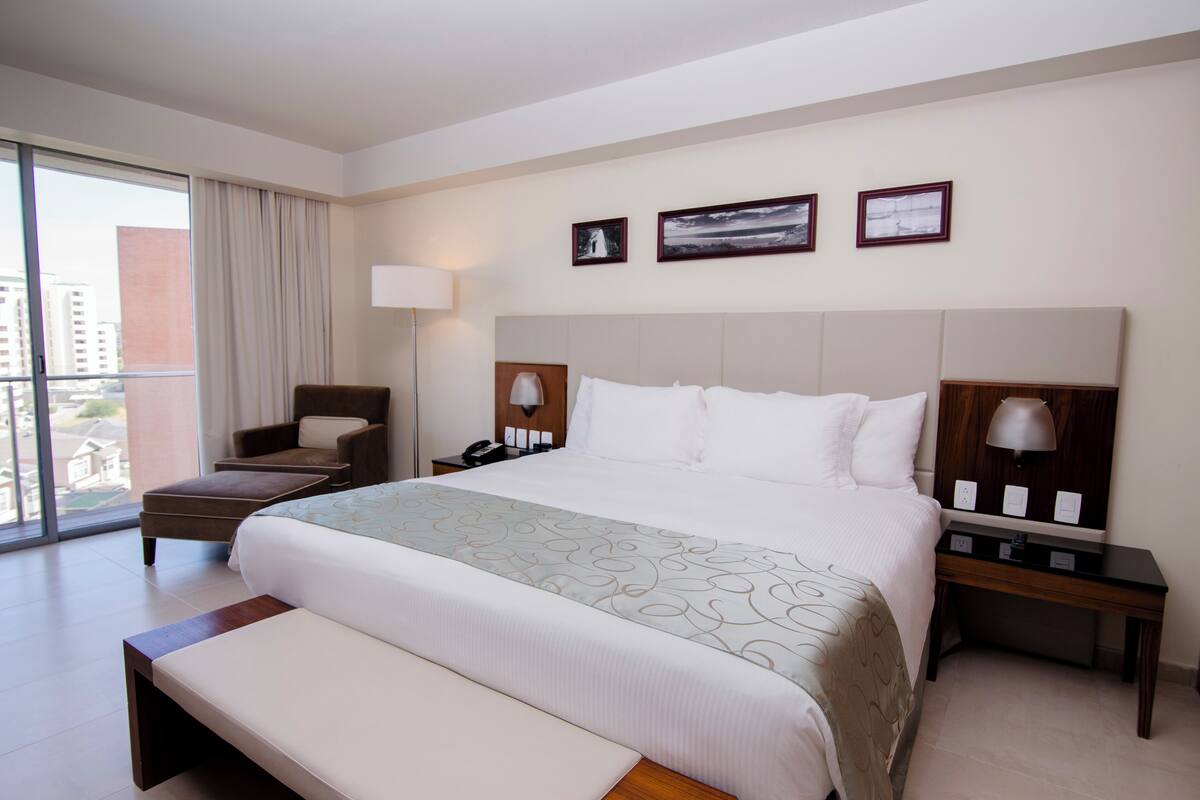Une chambre d’hôtel avec un grand lit, une chaise, une table de chevet, une lampe et une vue sur les bâtiments par la fenêtre.