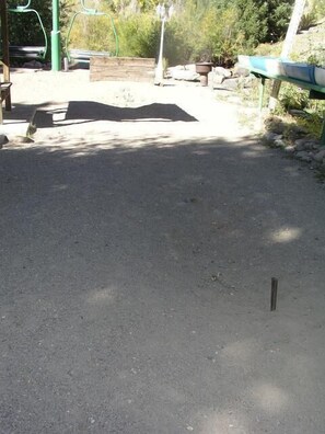 A paved area with a bench and a small table, surrounded by greenery and a wooden structure.