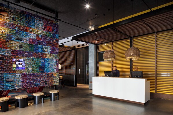 A modern reception area with a colorful mosaic wall, a white reception desk, and a seating area with stools. - Alt Hotel Winnipeg (Winnipeg)