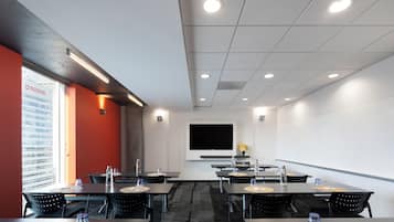 Un salón de conferencias con mesas, sillas, una pantalla grande y un ventanal con vistas a edificios.