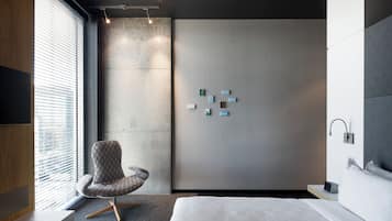 Una habitación de hotel moderna con una cama grande, un área de descanso con un sillón estampado, una lámpara fijada a la pared y una mesita con un control remoto.