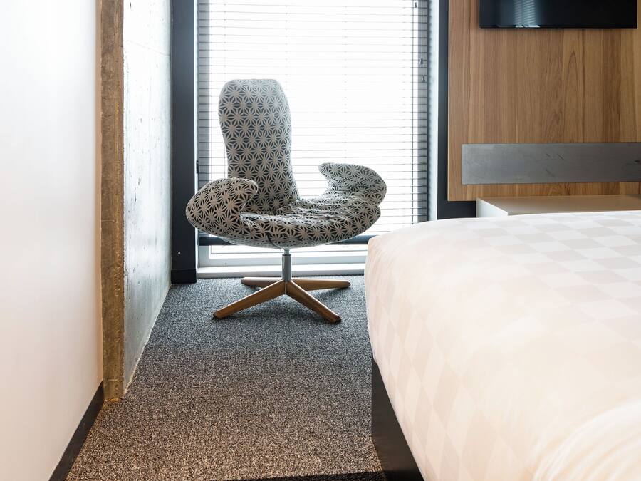 A modern hotel room with a large bed, a wooden headboard, a TV mounted on the wall, a patterned armchair, and a window with blinds.