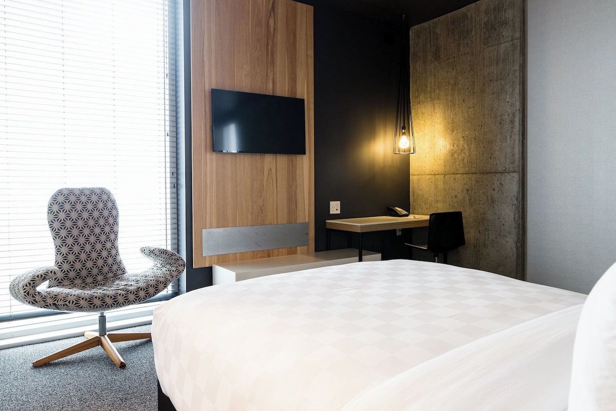 Ein modernes Hotelzimmer mit einem großen Bett, einer Wandpaneele aus Holz, einem Wand-Fernseher, einem Schreibtisch mit Stuhl und einer Pendelleuchte.