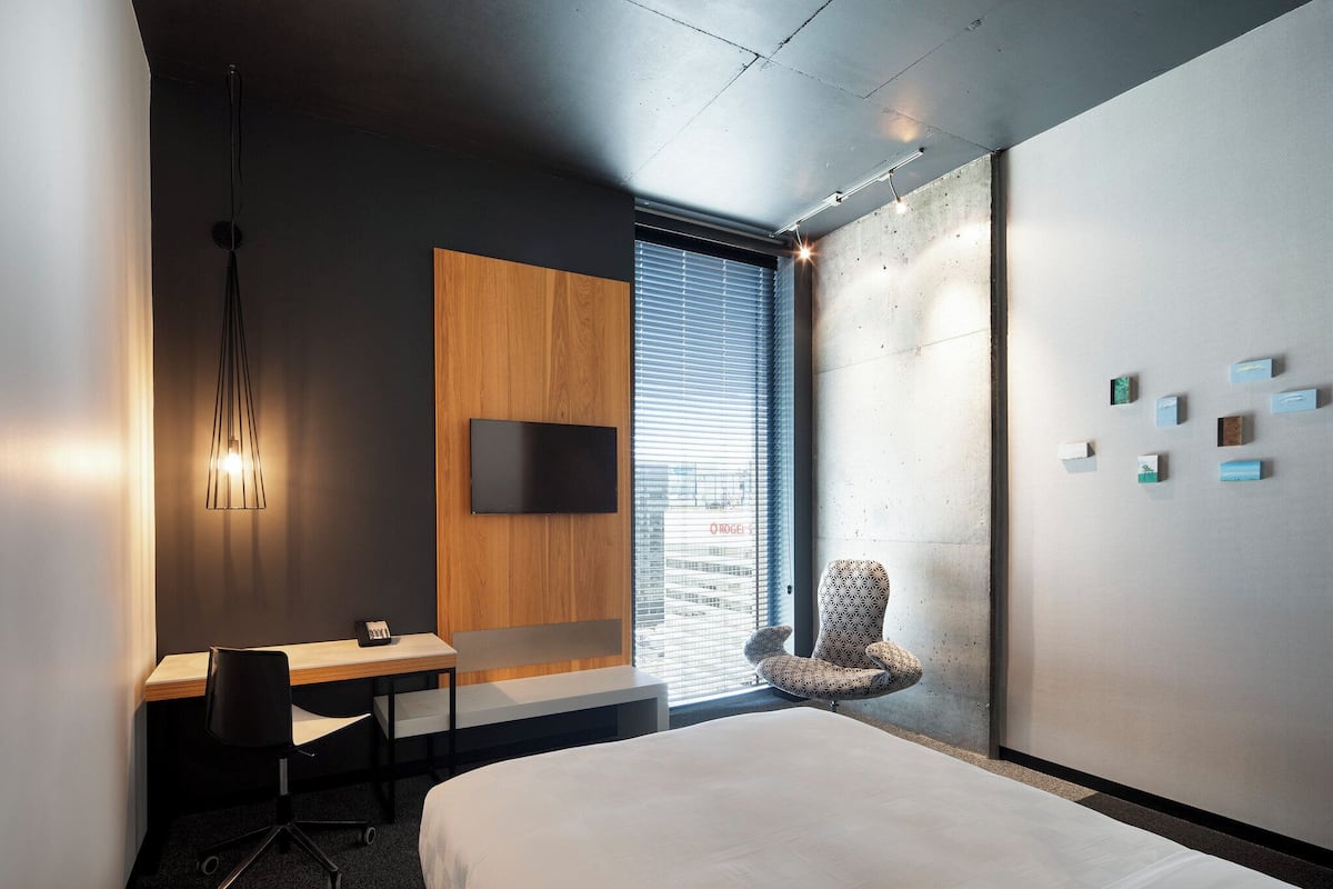 Ein modernes Hotelzimmer mit einem großen Bett, einem Schreibtisch mit Stuhl, einem an der Wand befestigten Fernseher und einem Fenster mit Jalousien.