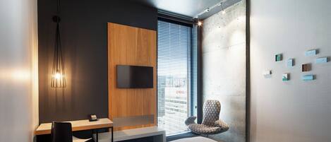 Habitación de hotel moderna con una cama grande, un escritorio con silla, un televisor montado en la pared y una ventana con persianas.