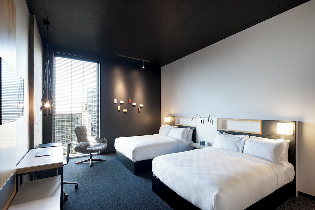 Ein Hotelzimmer mit zwei Betten, einem Schreibtisch, einem Stuhl und einem großen Fenster mit Jalousien.