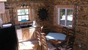 A rustic cabin interior with a wooden dining table, chairs, a kitchen area with a sink and cabinets, and a window providing natural light.