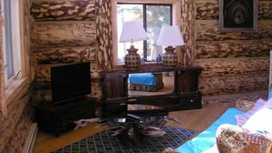 A rustic cabin interior with a wooden wall, a television on a stand, a desk with two lamps, and a patterned rug.