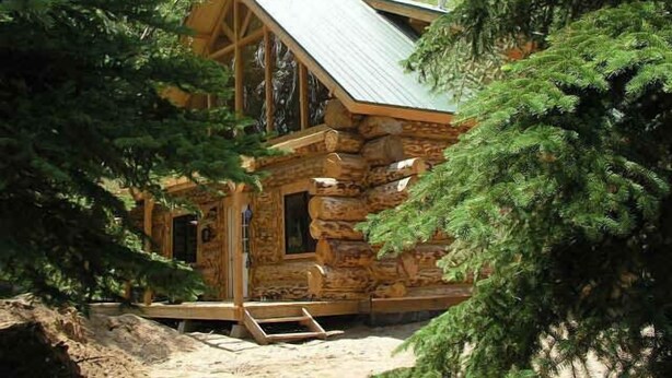 A rustic log cabin with a steeply pitched roof, surrounded by dense forest.