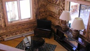 A log cabin interior with a television, two lamps, a coffee table, and a window providing a view outside.