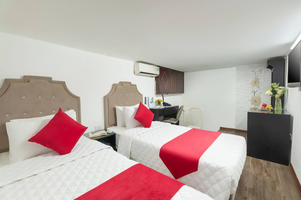 A hotel room with two beds, each with white bedding and red accents, a desk with a chair, a TV, and a small table with a vase of flowers.