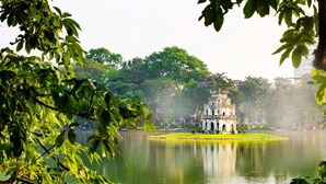 Un parque sereno con una estructura central similar a un pagodas, rodeado de un lago tranquilo, abundante vegetación y una ciudad distante.