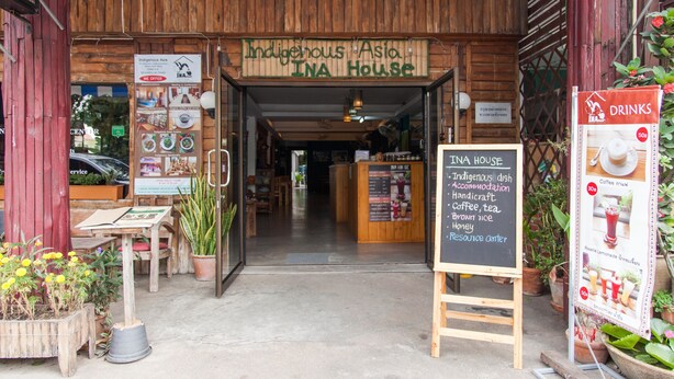 The entrance to Indigenous Asia INA House with a wooden facade, a signboard listing amenities like coffee, tea, and brown rice, and a small seating area with potted plants. - INA House (Chiang Mai)