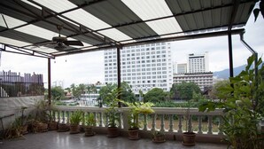 A covered balcony with potted plants, a metal railing, and a view of tall buildings in the background. - INA House (Chiang Mai)