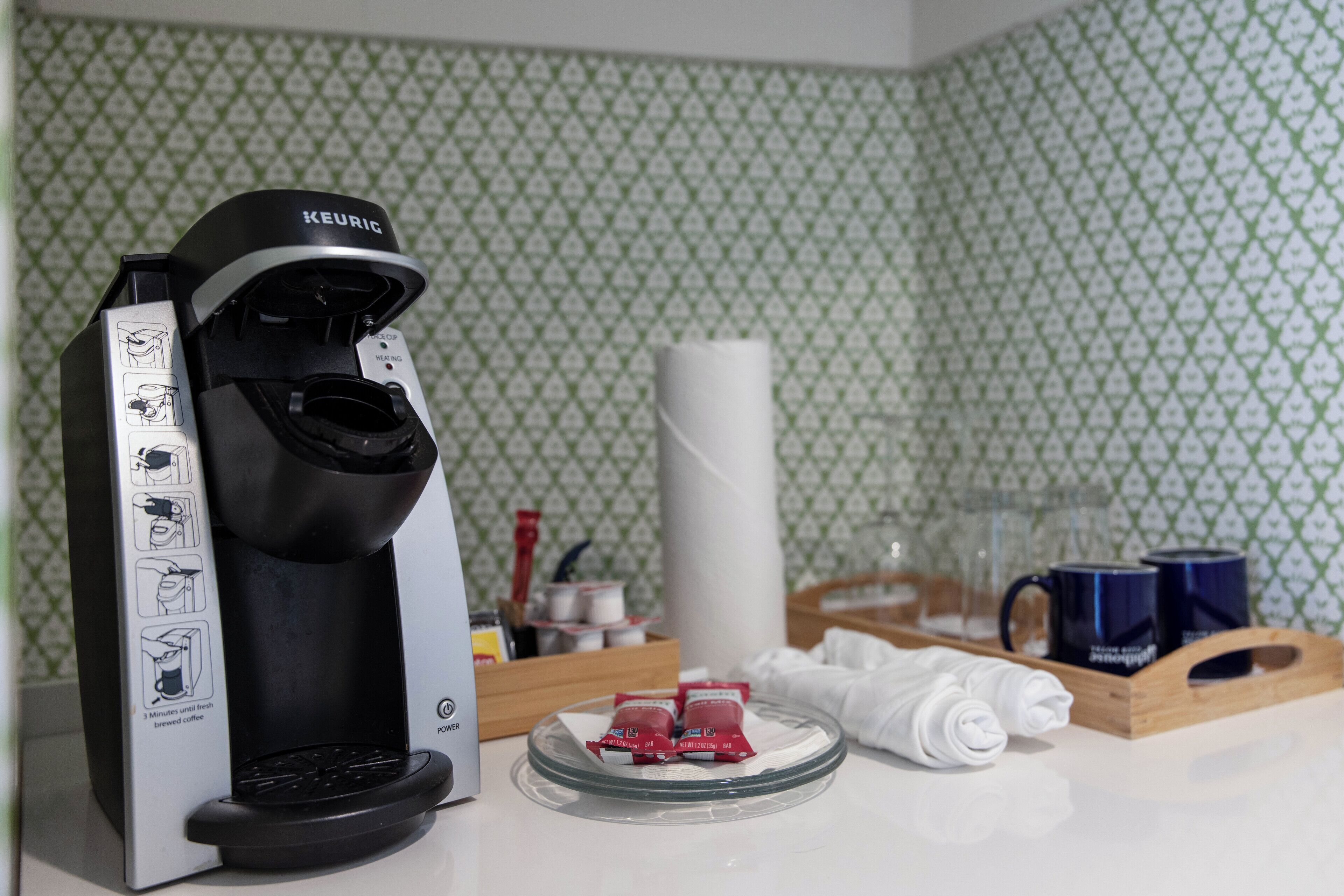 Una cafetera Keurig, un plato de vidrio con cápsulas de café, un rollo de toallas de papel y una bandeja con tazas y un vaso.