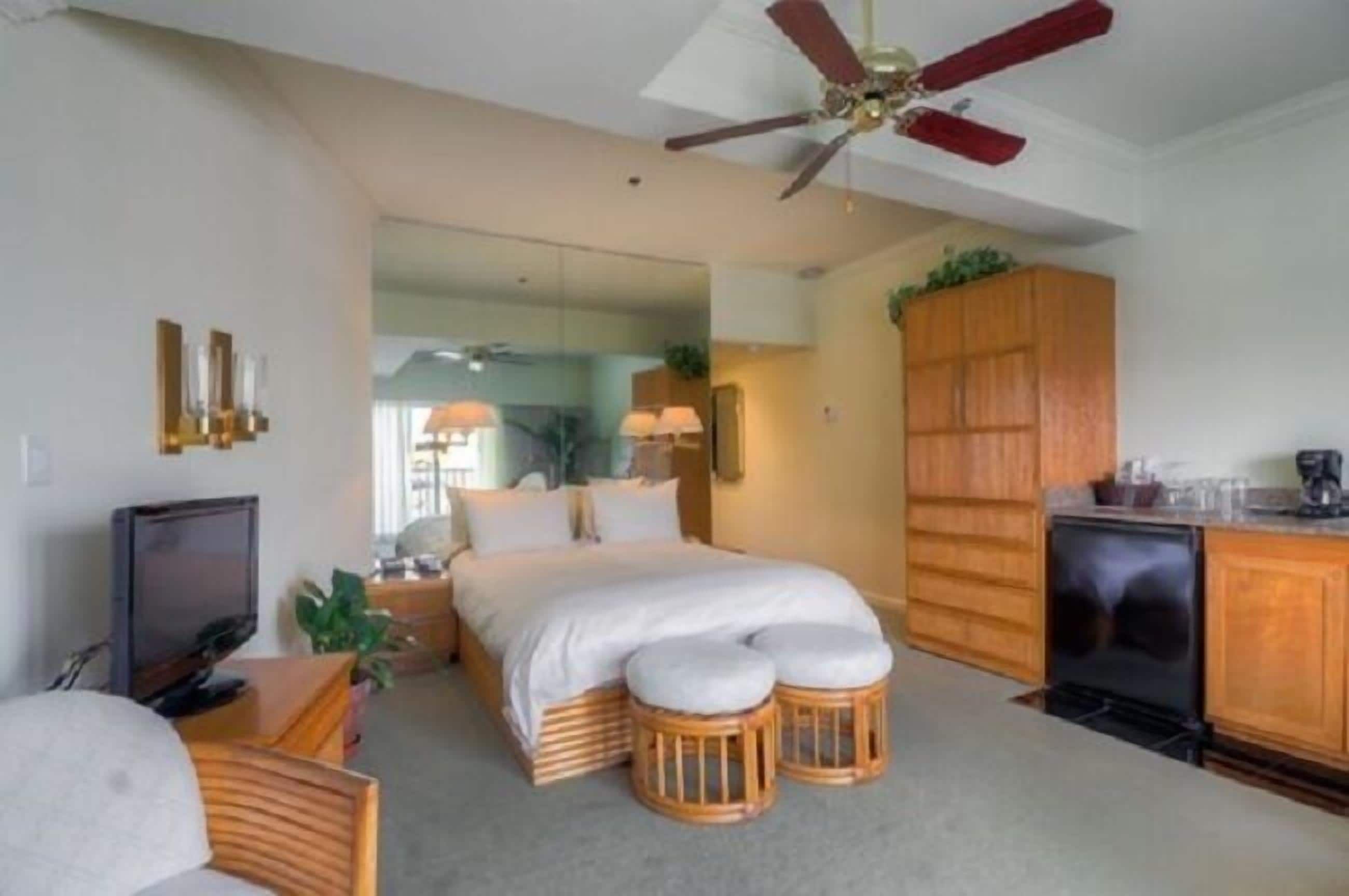 A bedroom with a bed, bedside tables, a television, a ceiling fan, and a kitchen area with cabinets and a refrigerator.