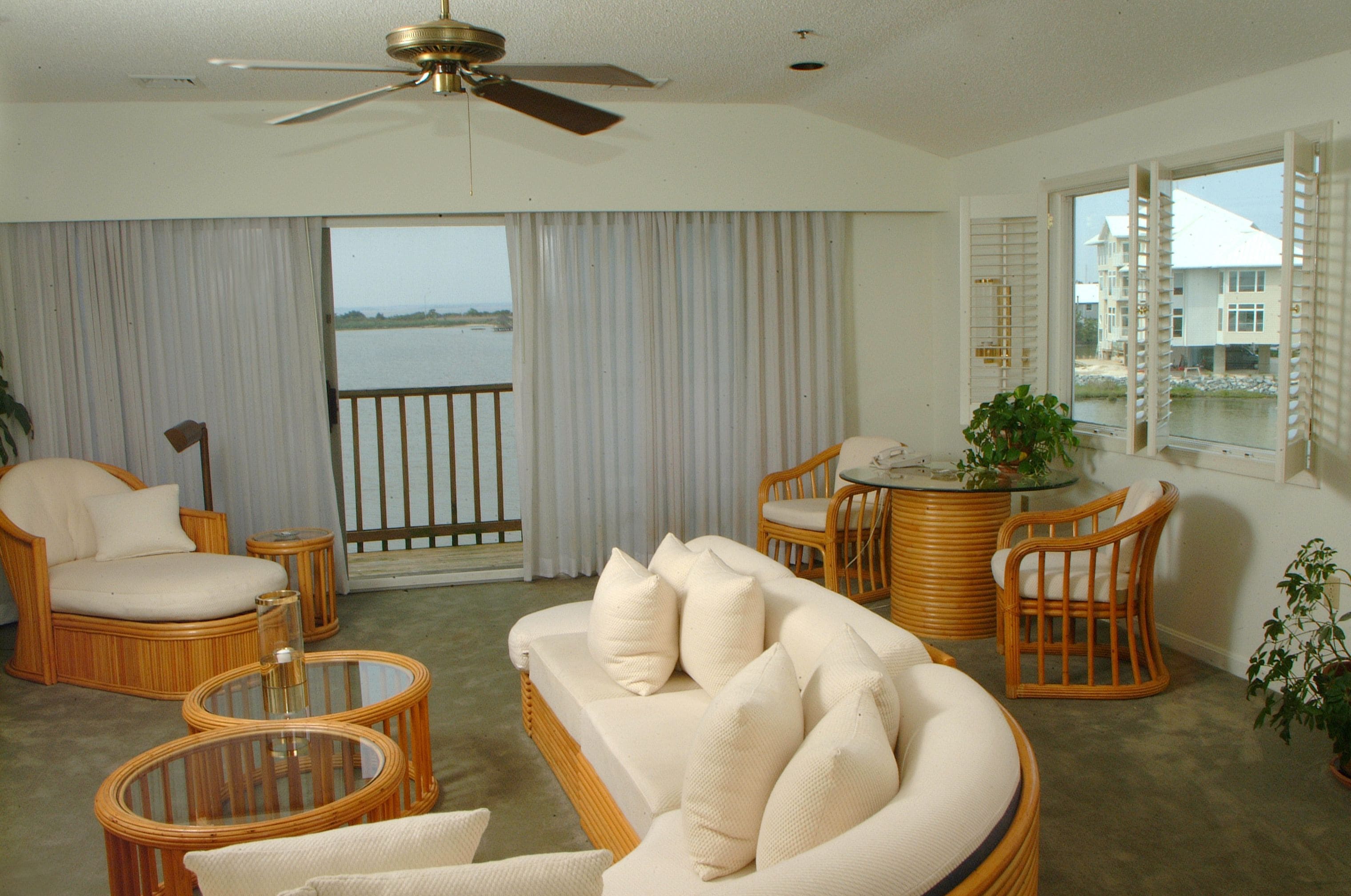 A spacious living room with a large sofa, two armchairs, and a coffee table. A ceiling fan is visible, and the room has a view of a body of water through large windows.