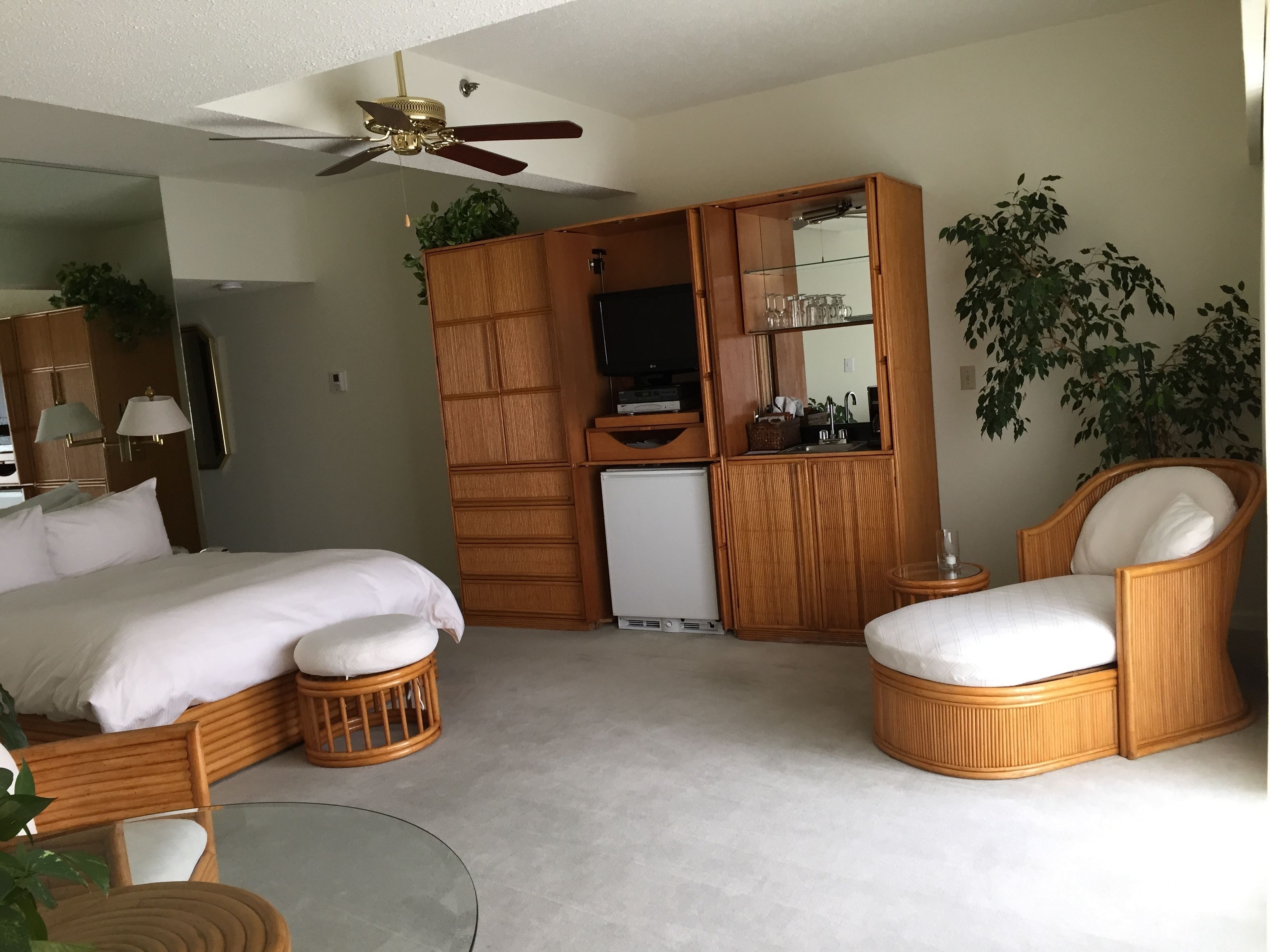 Une chambre à coucher comprenant un lit, deux fauteuils en osier, une petite table, un ventilateur de plafond, une armoire, un miroir et une plante en pot.