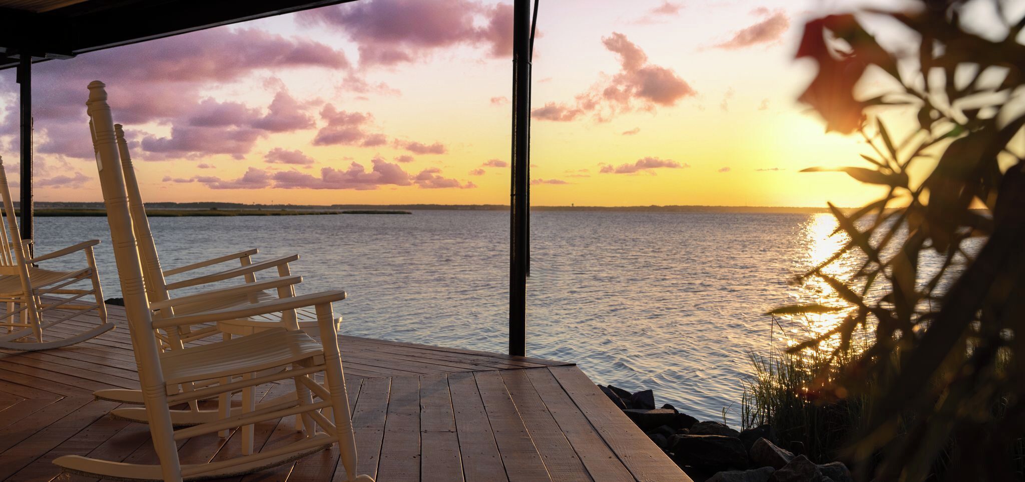 Un muelle de madera con mecedoras dando vista a un cuerpo de agua al atardecer.