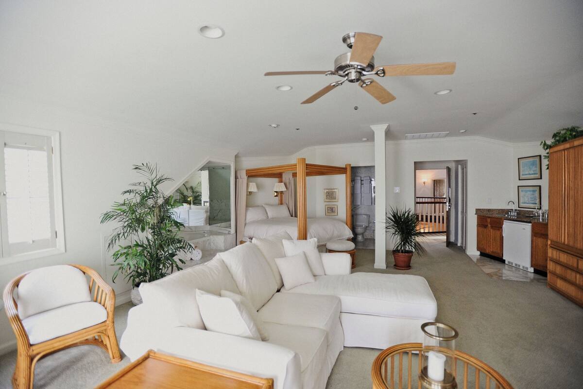 A spacious living room with a large white sectional sofa, a wooden chair, and a ceiling fan. There is a kitchen area visible in the background.
