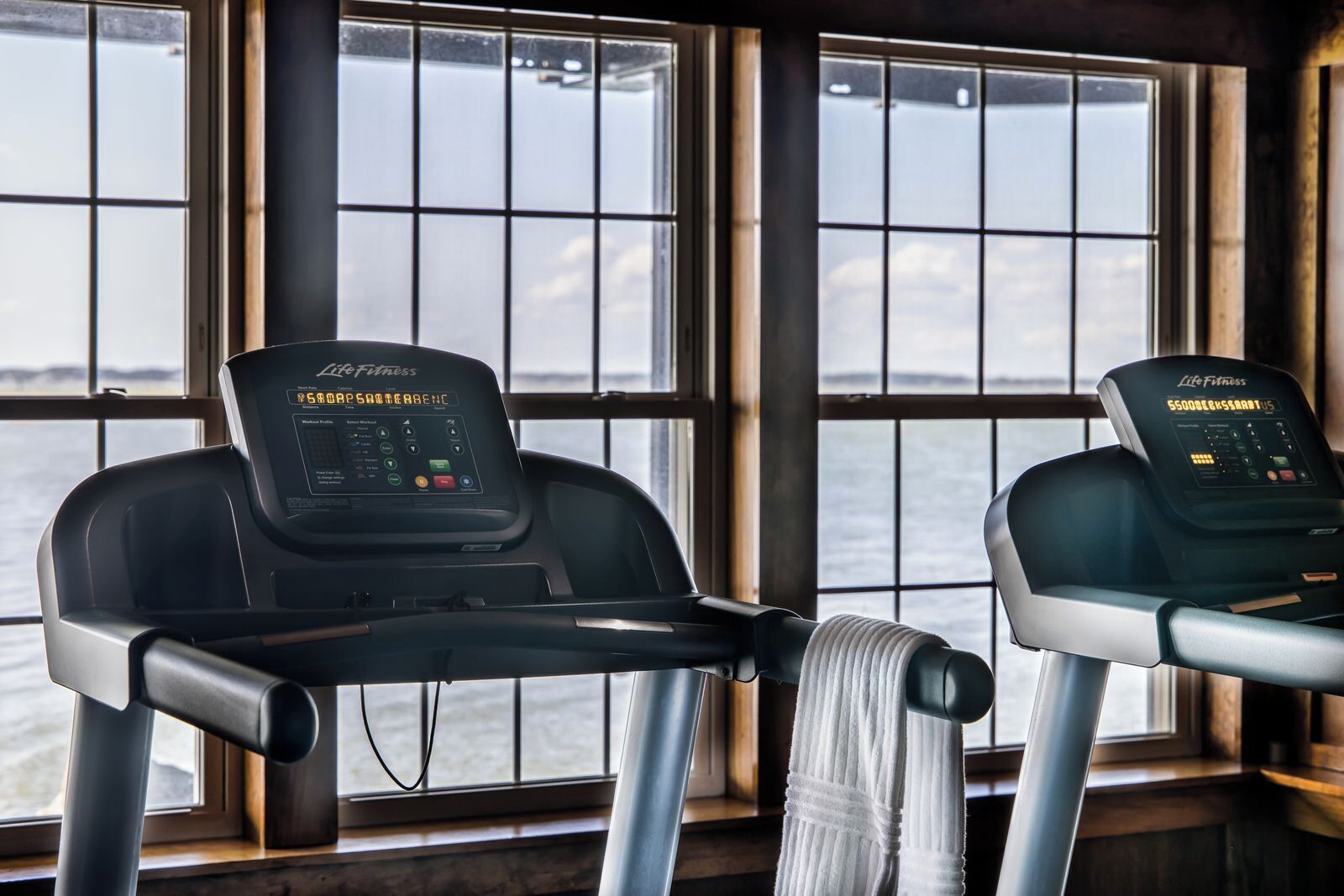 Two treadmills with digital displays in a room with large windows overlooking a body of water.