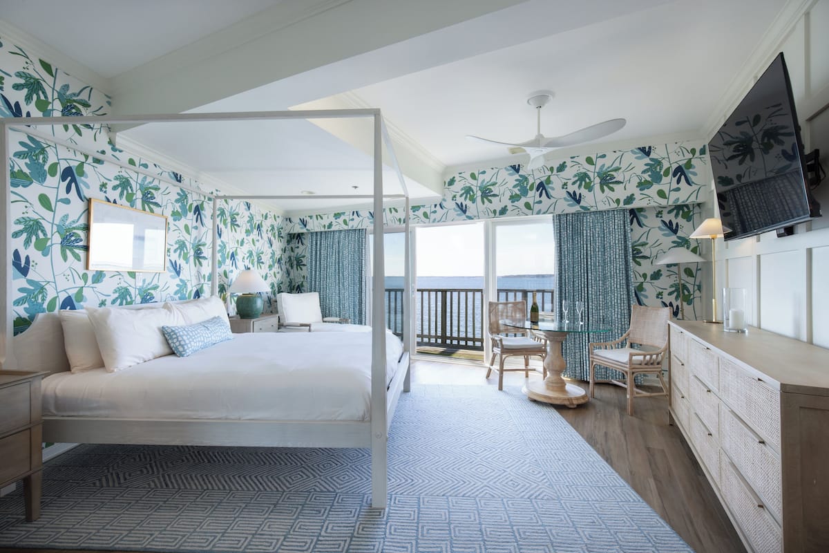 A bedroom with a bed, a desk, and a chair, a balcony with a view, and a television on the wall.
