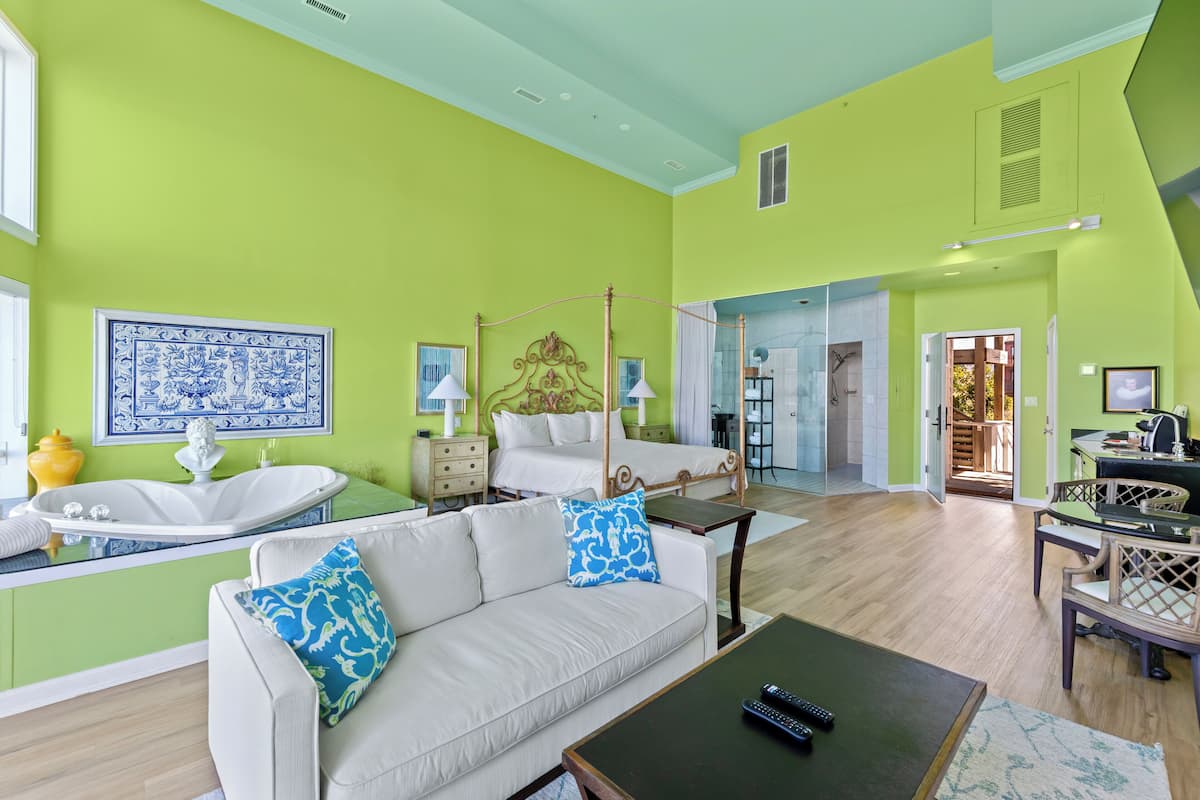 A spacious living area with a large sofa, a coffee table, and a dining table with chairs. There is a large window providing natural light, and a bathroom with a bathtub and sink is visible in the background.