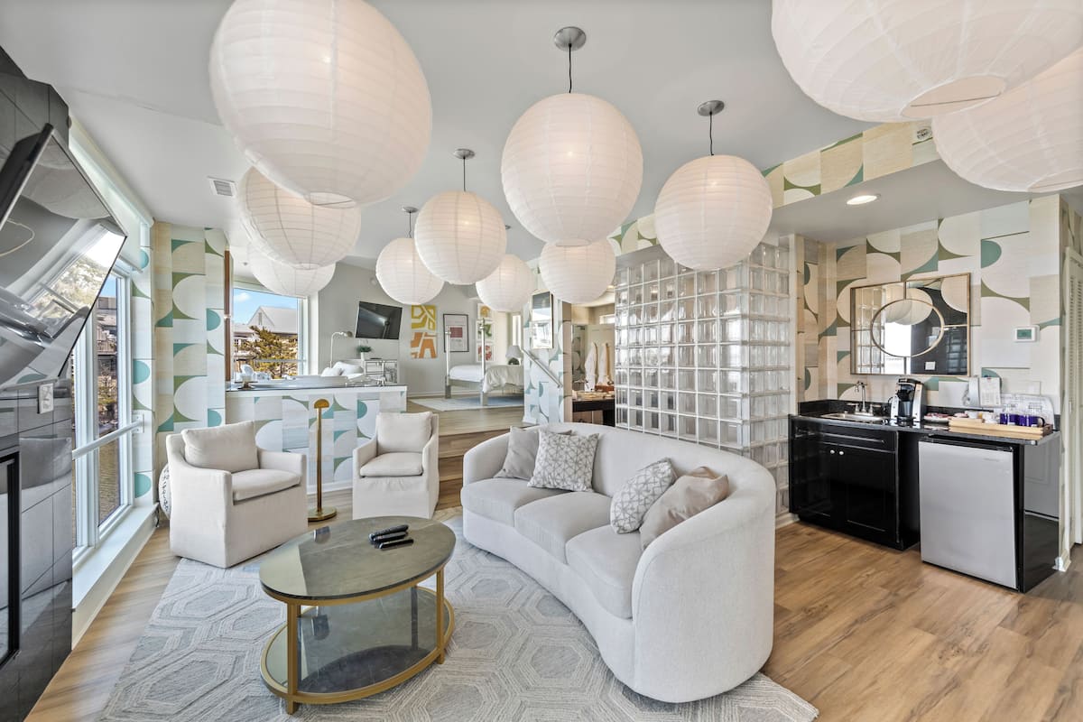 A modern living room with a sofa, armchairs, and a coffee table. There are large windows and a kitchen area visible.