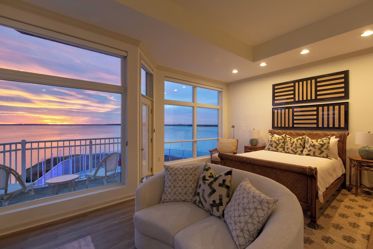 A bedroom with a large window overlooking a body of water at sunset, a bed with patterned pillows, a chair, and a nightstand with a lamp.