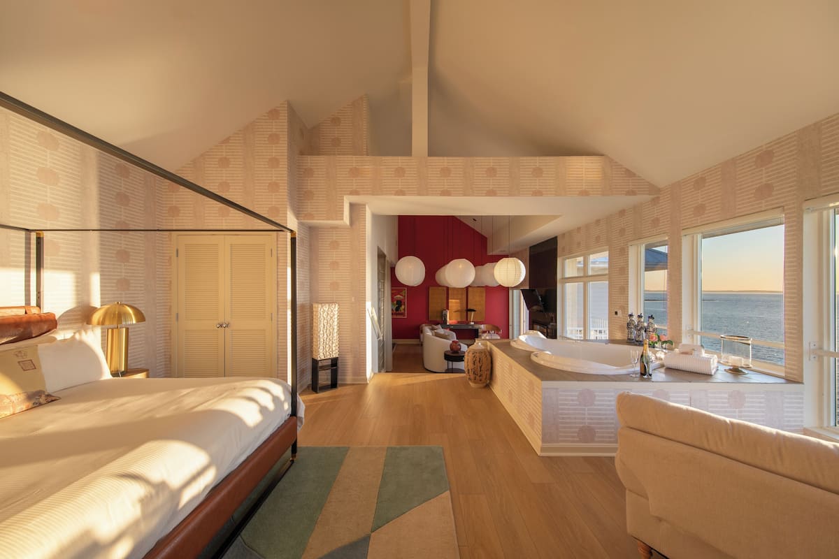 A spacious bedroom with a large bed, a sitting area with a view of the sea, and a separate bathroom with a bathtub.
