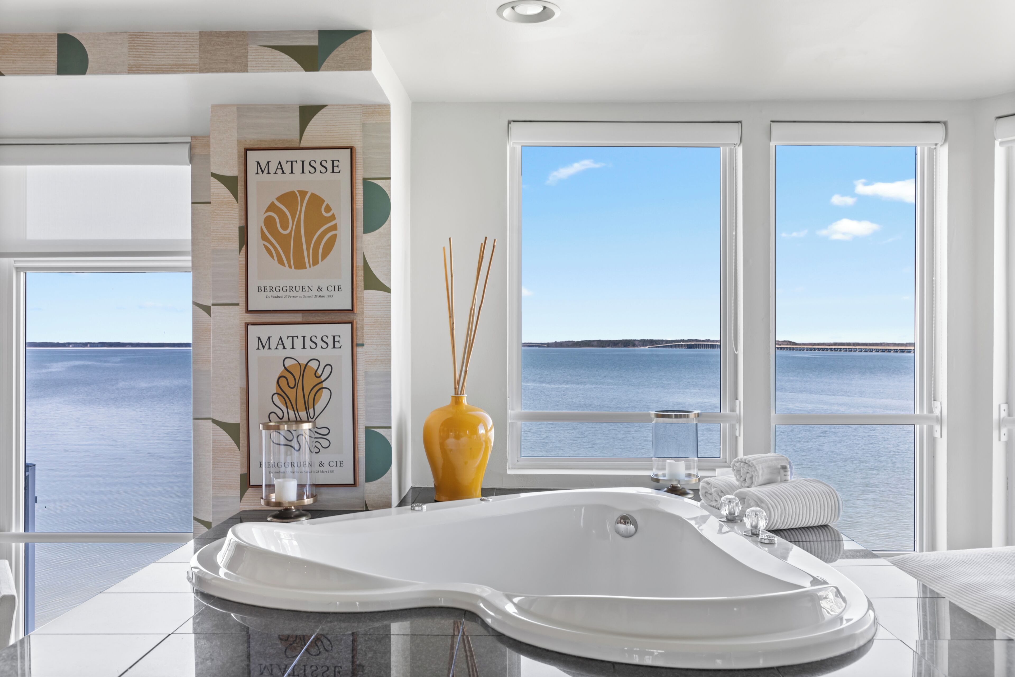 A modern bathroom with a large bathtub, two framed artworks, a vase, and a view of the sea.