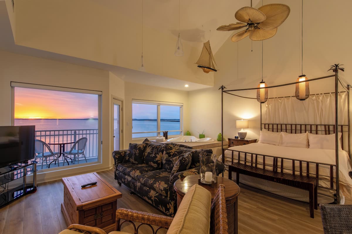 A bedroom with a bed, bedside table, lamp, and a view of the sea through a large window.