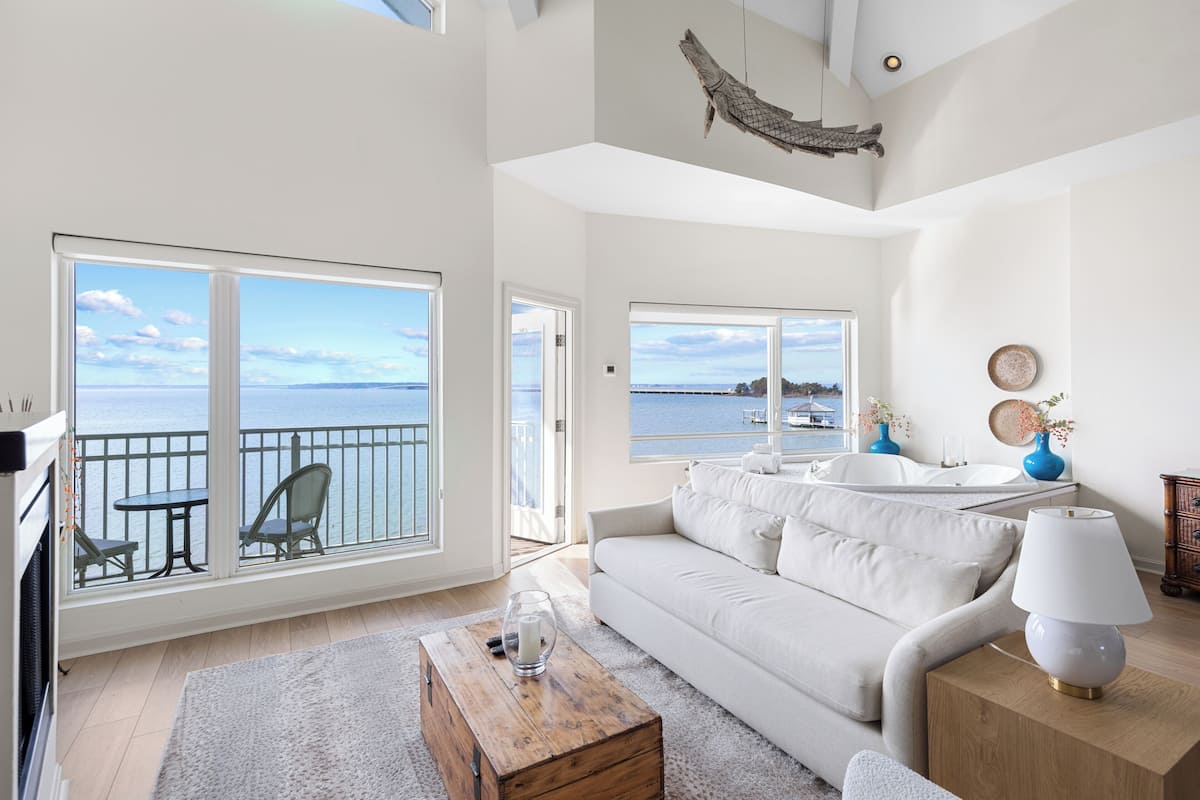 A spacious living room with a large window overlooking the sea, a white sofa, a wooden coffee table, and a lamp.