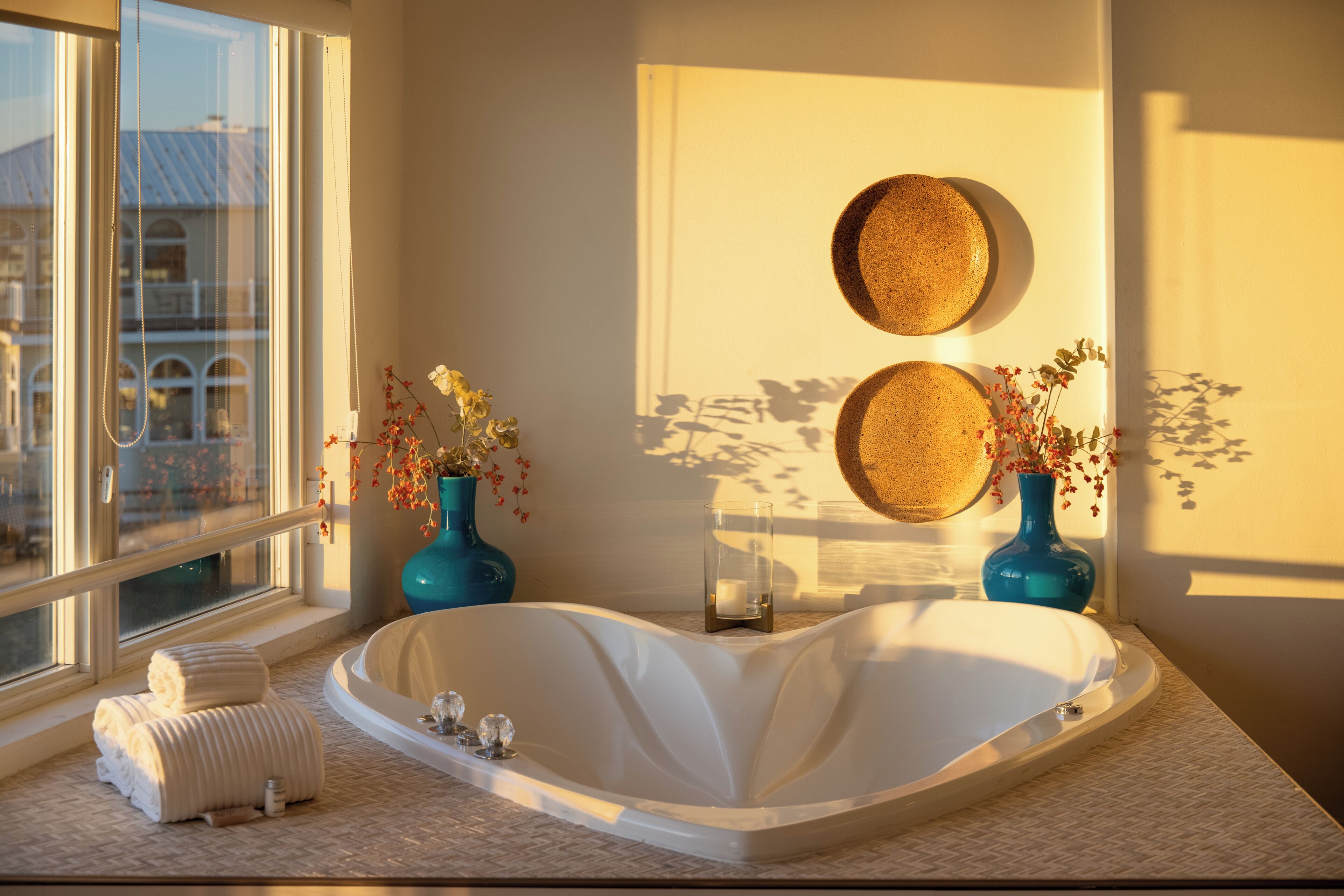 A modern bathroom with a freestanding bathtub, two vases with flowers, and a window with a view of buildings.