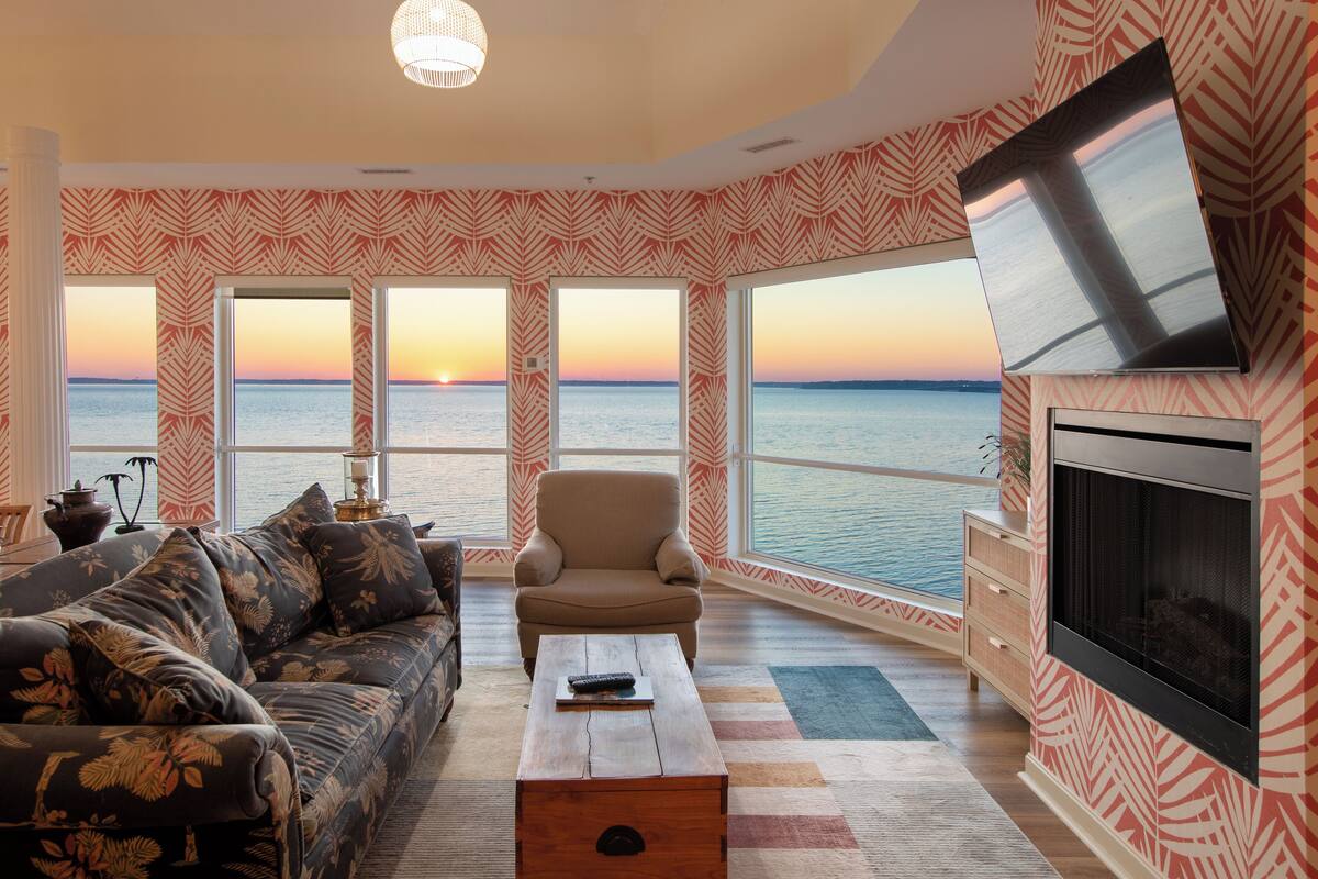 A living room with a sofa, armchair, coffee table, and a TV mounted on the wall. Large windows provide a view of the sea.