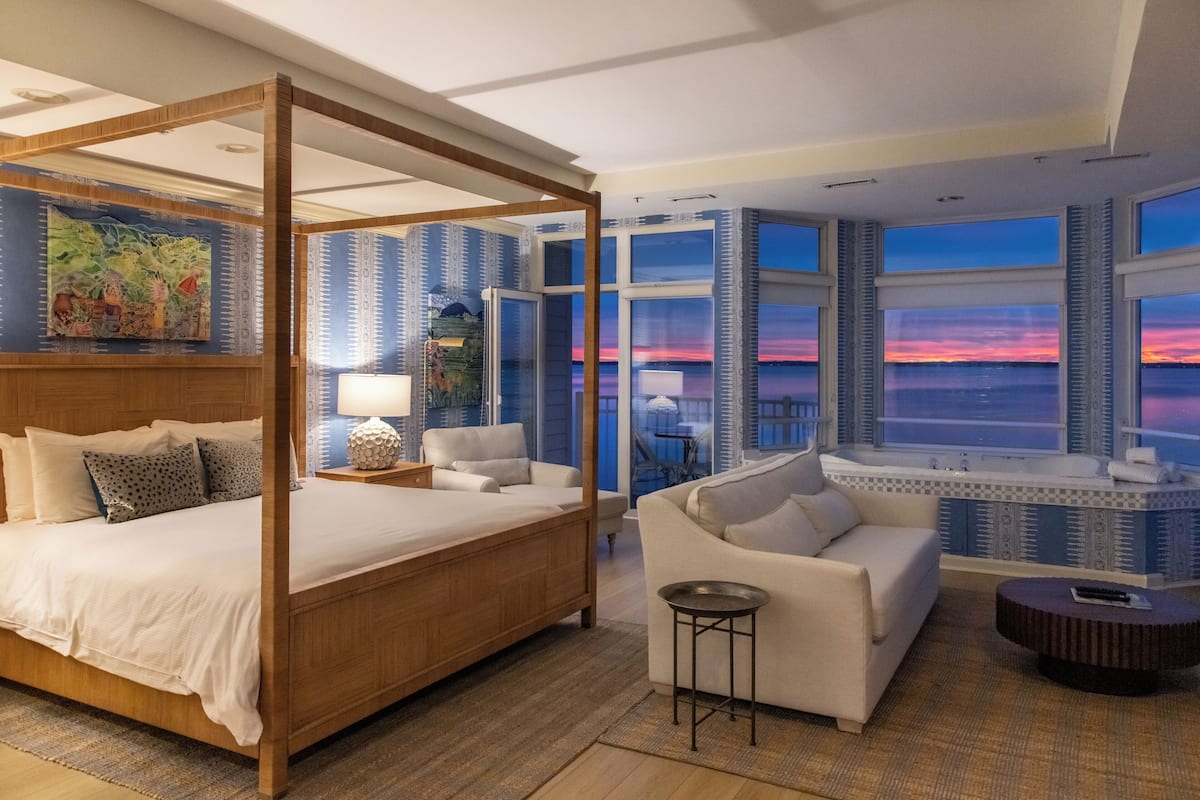 A spacious bedroom with a four-poster bed, a sitting area with a sofa and armchairs, and a view of the sunset through large windows.