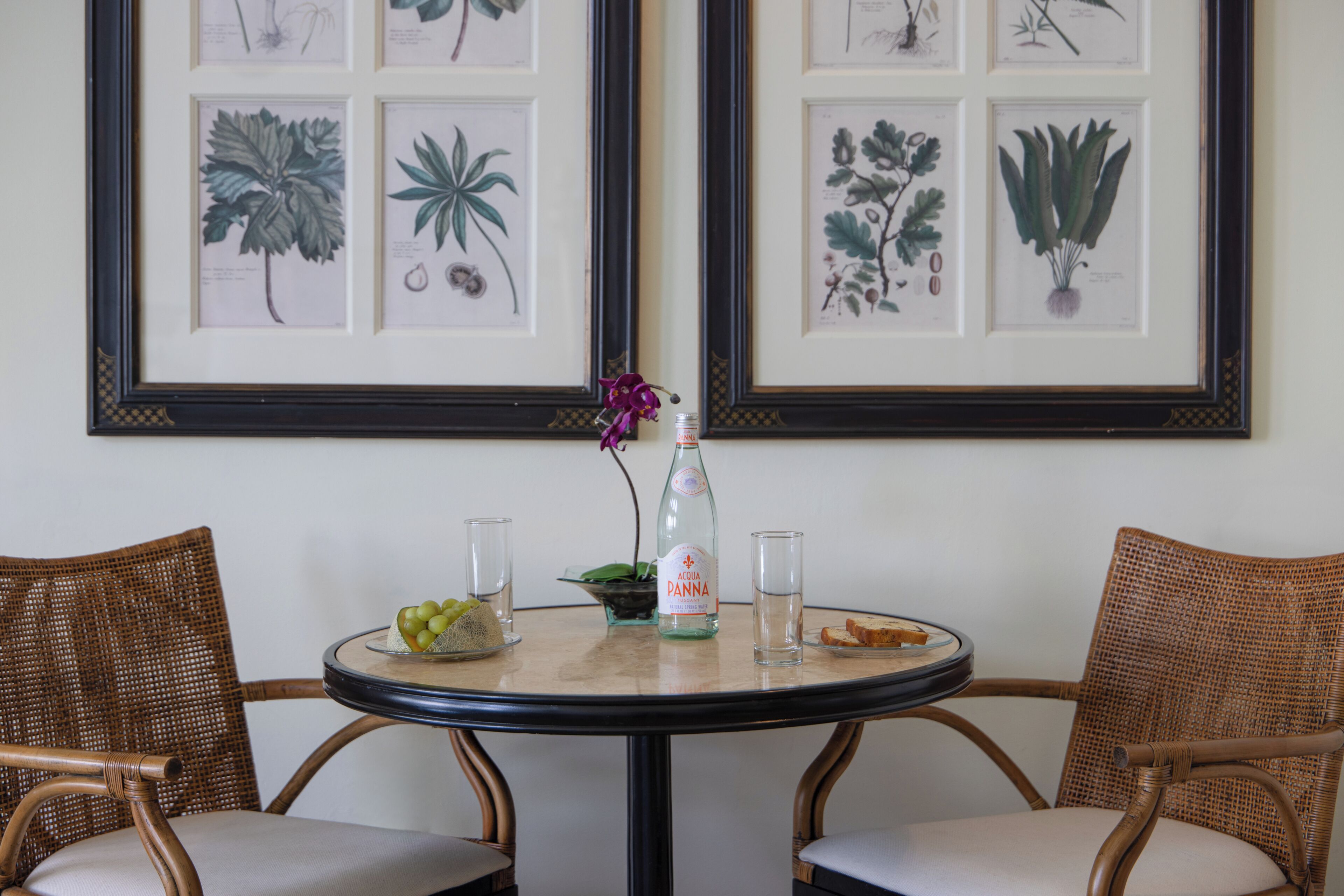 A round table with chairs, a bottle of Panna, a glass, and a bowl of fruit.