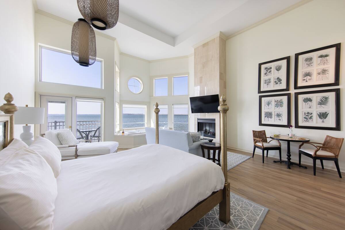 A spacious bedroom with a large bed, a sitting area with a view of the ocean, a fireplace, and framed botanical prints on the wall.
