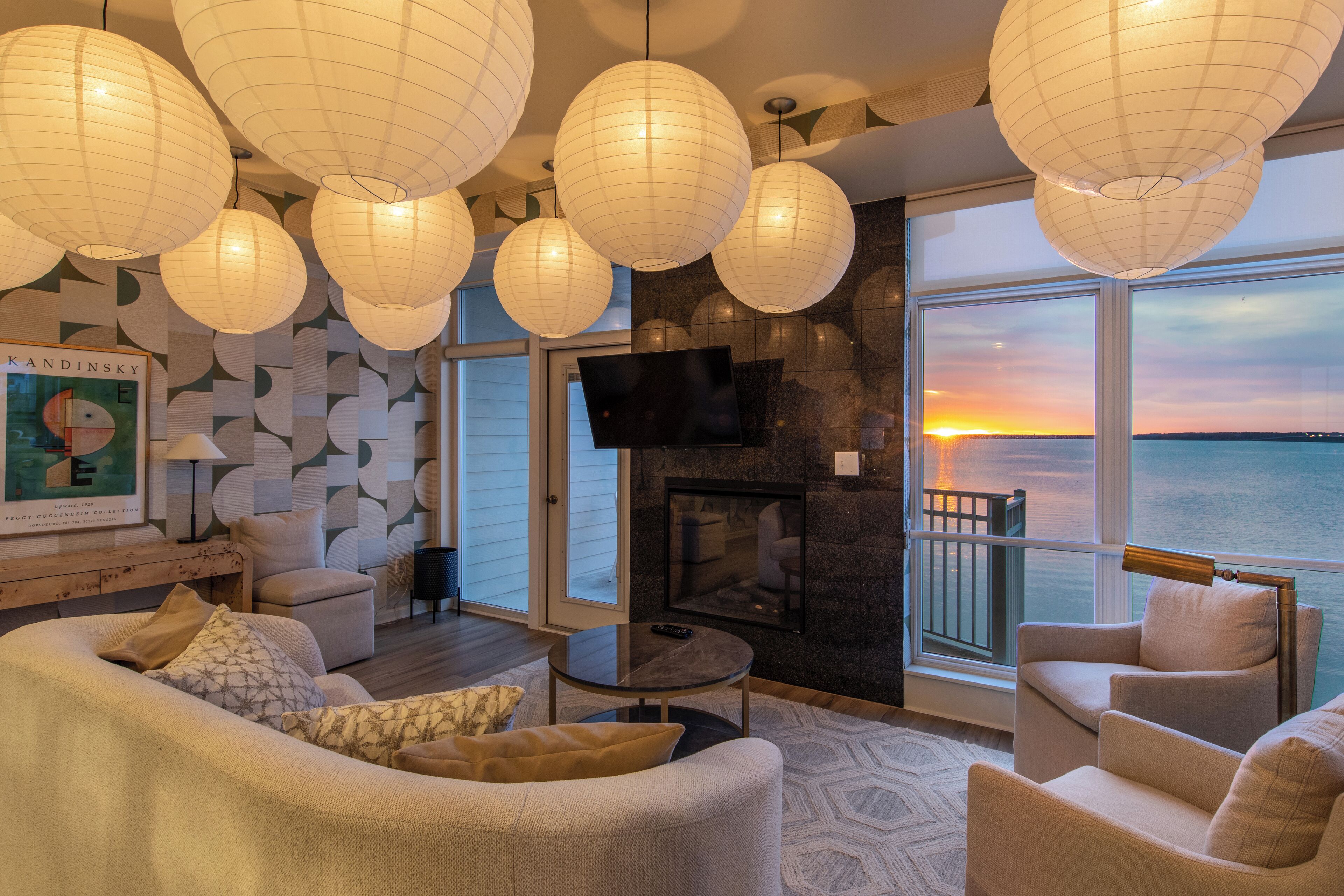 A modern living room with a fireplace, comfortable seating, and large windows overlooking the ocean at sunset.