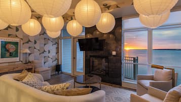 A modern living room with a fireplace, comfortable seating, and large windows overlooking the ocean at sunset.