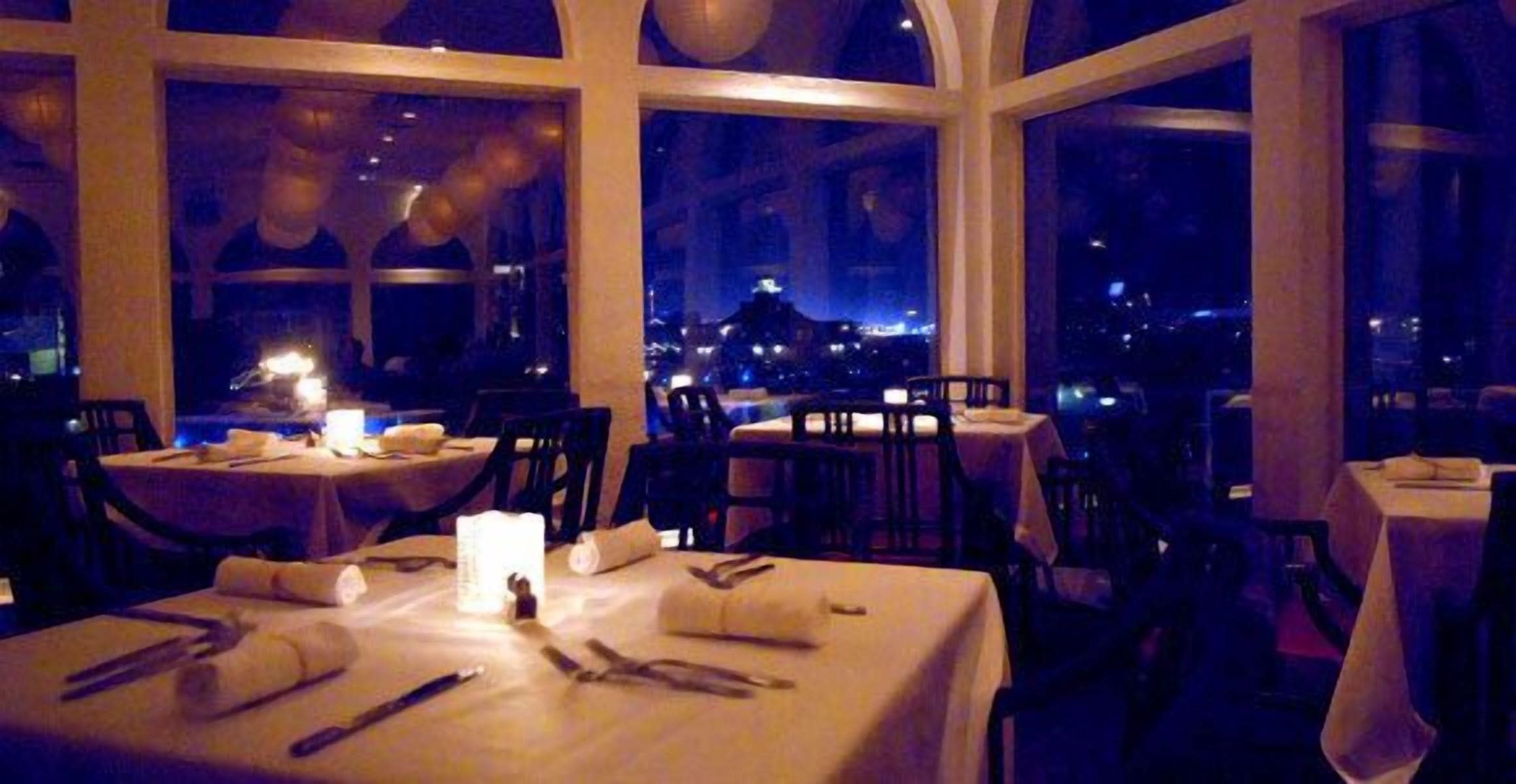 Un restaurante con mesas dispuestas para comer, ventanas amplias y vista nocturna a la ciudad.