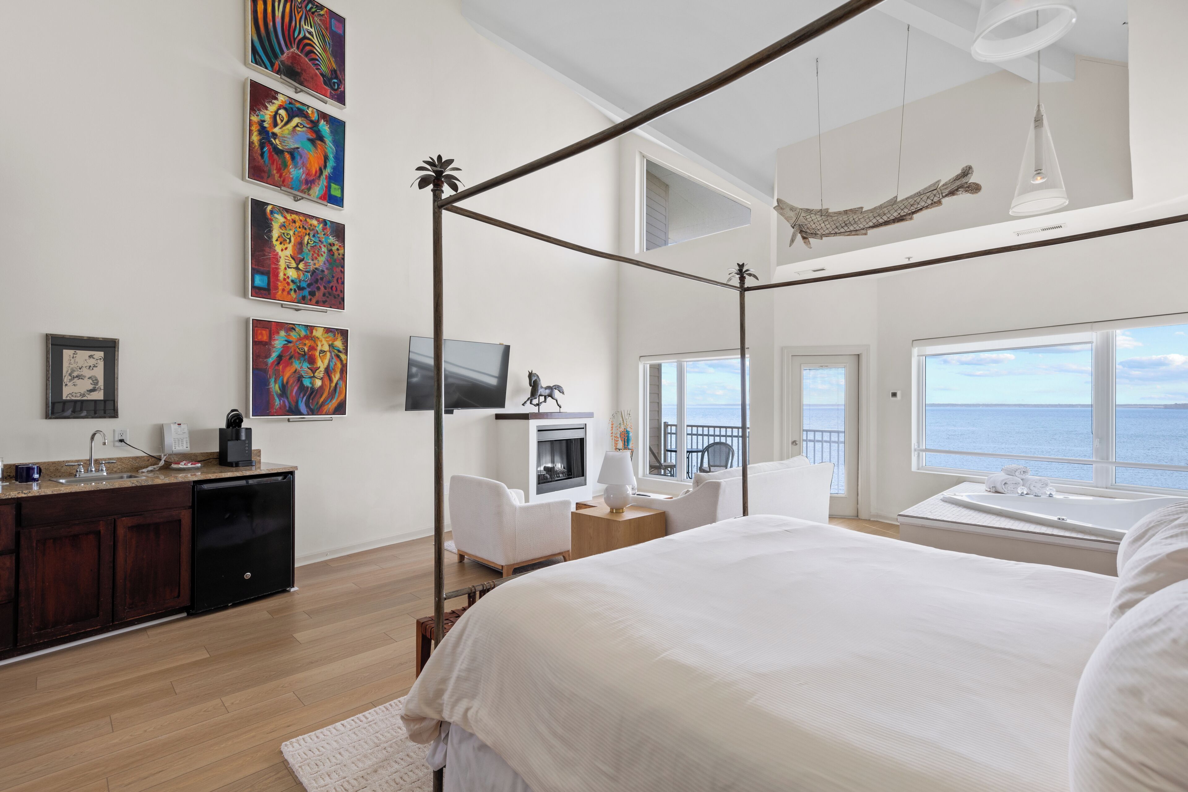 A bedroom with a four-poster bed, a television, a fireplace, a balcony with a view of the ocean, and artwork on the wall.