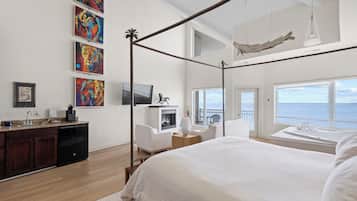 A bedroom with a four-poster bed, a television, a fireplace, a balcony with a view of the ocean, and artwork on the wall.